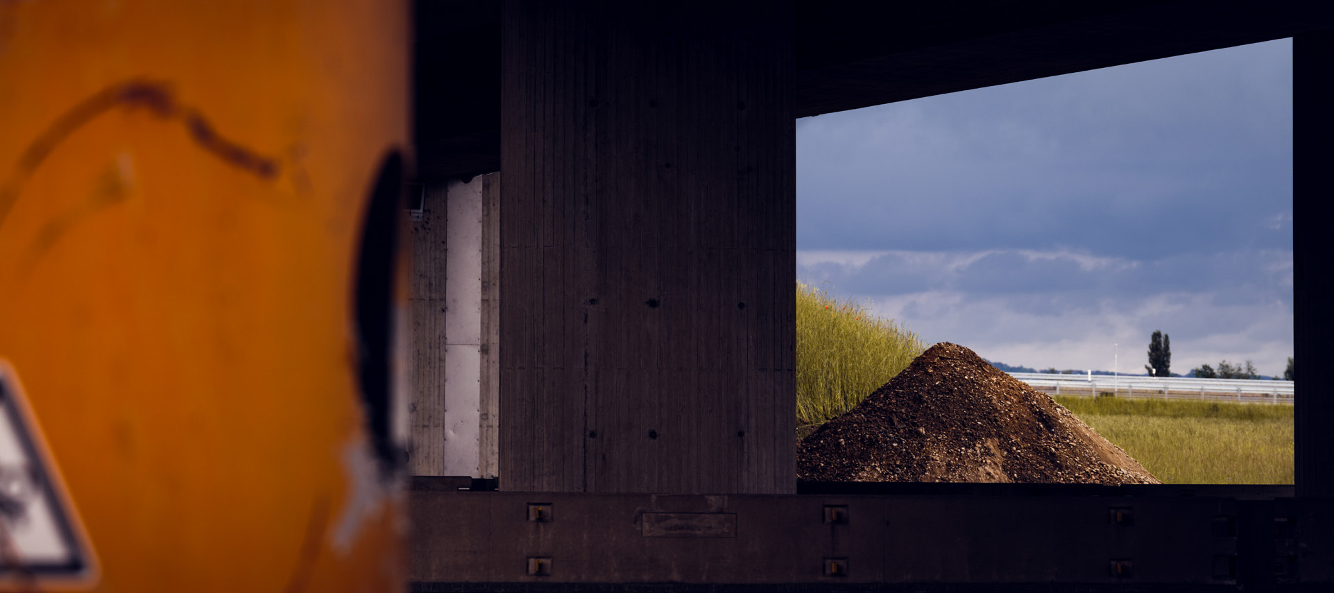 Das Bild zeigt einen Blick durch eine Betonbrücke oder ein großes Bauwerk auf einen Erdhügel und eine ländliche Landschaft.